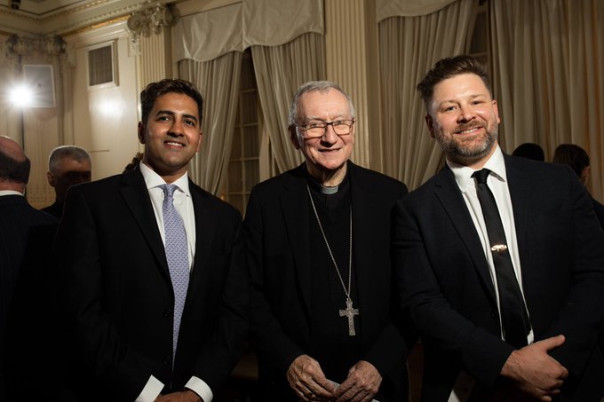 Three men in formal attire pose for a photograph in an elegant room with classical architecture; the central figure wears a clerical collar and a large cross necklace.