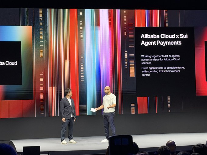 Two men converse on stage in front of a large screen displaying "Alibaba Cloud x Sui Agent Payments," with text explaining the partnership to let AI agents access and pay for Alibaba Cloud services while giving owners spending controls.