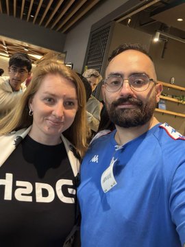 A selfie photo of a smiling woman and a bearded man in glasses, taken indoors with a lanyard visible around the man's neck.
