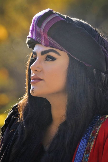 A close-up portrait of a woman wearing a dark headscarf with purple accents, looking off to the side in warm, golden lighting.