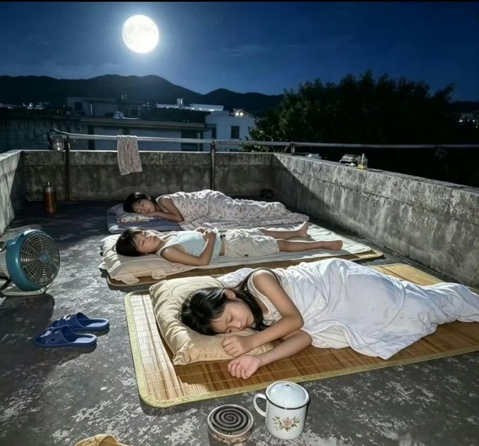 Three people in pajamas sleep on traditional mats on a rooftop at night under a bright full moon, with a fan, slippers, and a mug nearby.