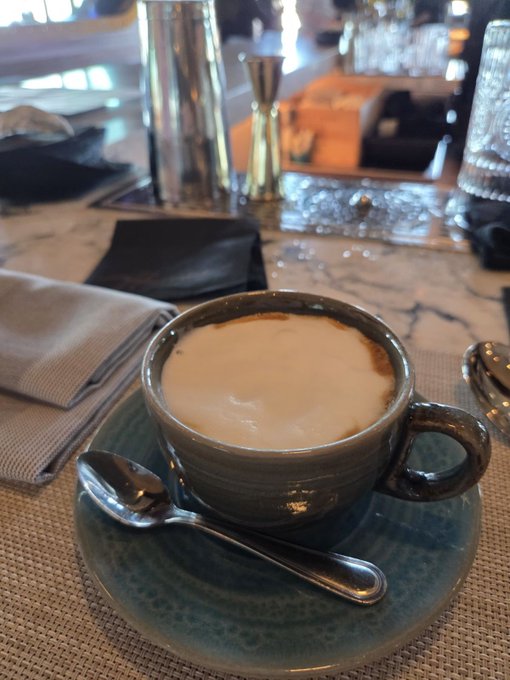 A close-up shot features a cup of cappuccino in a blue ceramic cup and saucer, resting on a textured placemat with a silver spoon.