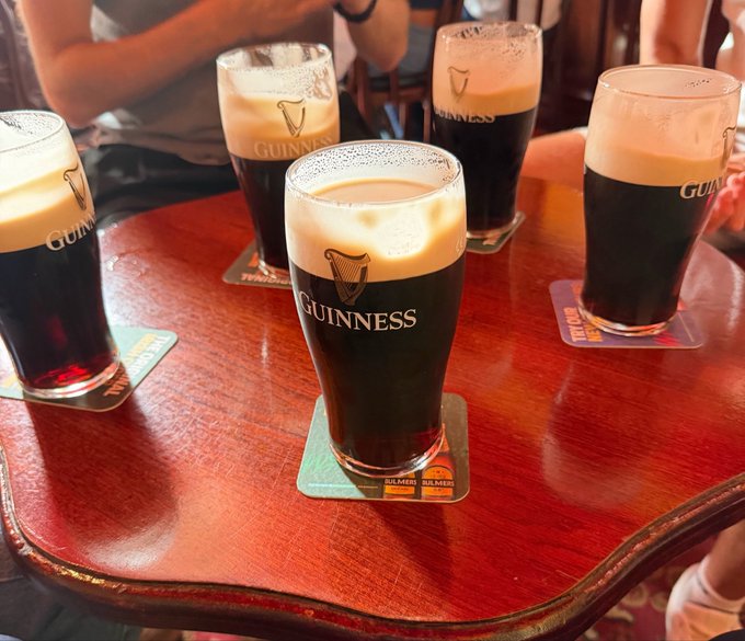 Four pints of dark Guinness stout with creamy heads sit on branded coasters atop a polished wooden pub table.