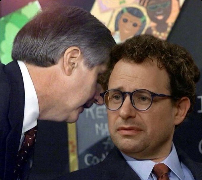 Two men in suits appear to be in a private conversation, with one man leaning in to whisper near the ear of the other man who wears round glasses and has a serious expression.