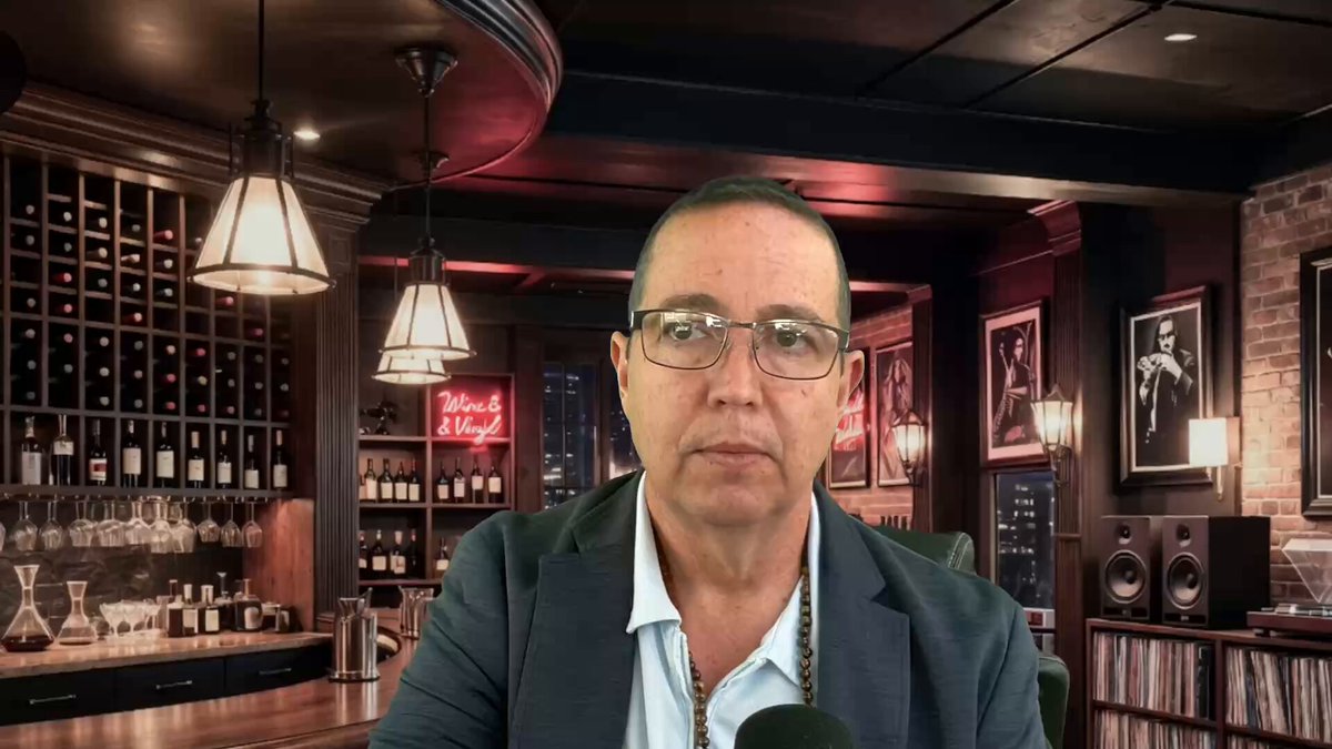 In the image, a man wearing glasses is sitting at a bar with several bottles of wine behind him. The setting appears to be an indoor space with a dark atmosphere, possibly a restaurant or a bar. There are multiple wine bottles on display, indicating that this could be a place where people gather for drinks and socializing. A microphone can also be seen in the scene, suggesting that there might be some form of live entertainment taking place at the venue. The post includes an image of a man wearing glasses sitting behind a bar with several bottles of wine displayed on the wall.