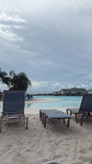 A sandy beach with two blue lounge chairs and a white lounger facing turquoise water and a resort building, with palm trees and a cloudy sky.