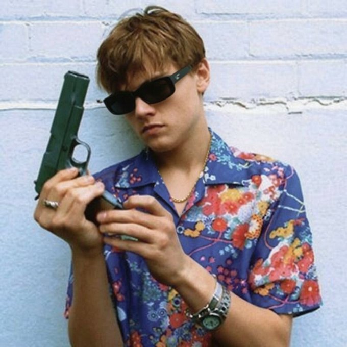 A young man wearing sunglasses and a colorful floral shirt holds a handgun while leaning against a wall, projecting a stylish yet edgy vibe that complements the bold, attention-grabbing tone of the memecoin giveaway promotion.

*(Note: I cannot identify the person in the image based on their facial features. If you could tell me who they are, I could provide more context about how the image relates to your post.)*