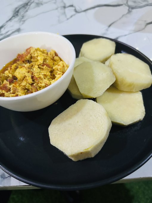 A meal served on a black plate over a marble-patterned table, featuring a white bowl filled with a yellow egg and tomato scramble alongside several thick slices of peeled boiled cassava or yam.