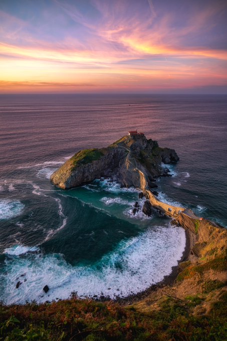 An aerial view of a rocky island with a stone bridge connecting to the mainland, topped with a lighthouse, set against a vibrant sunset sky and ocean waves.