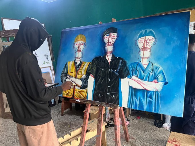 A person in a black hoodie works on a large painting of three masked figures with wireframe facial overlays, one in a yellow construction vest, one in a denim shirt, and one in blue scrubs.