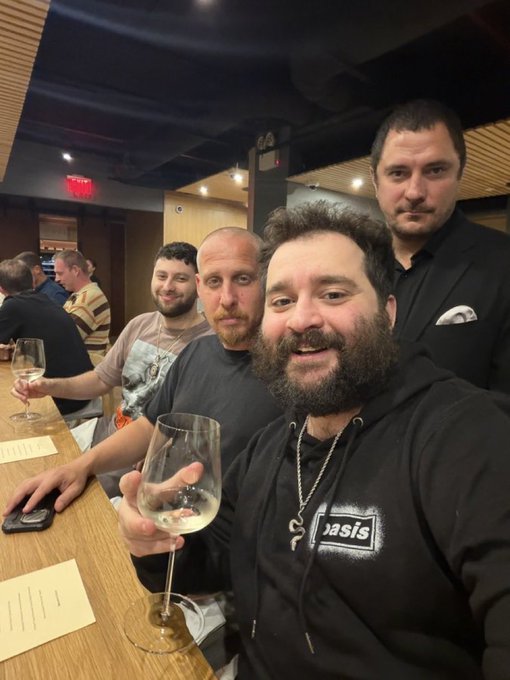 Group photo taken at a bar or restaurant featuring four men, with the man in front wearing an Oasis hoodie and holding a wine glass.
