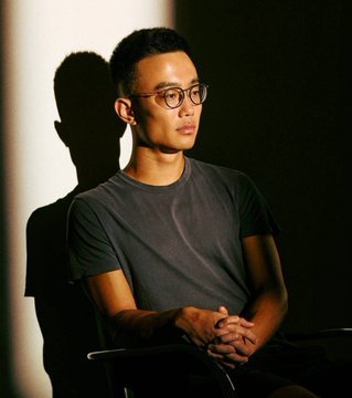 I cannot identify the specific person in this image based on their facial features. I can describe what I see: a young man with dark hair wearing glasses and a dark gray t-shirt, seated with his hands clasped together, lit dramatically with warm light from one side against a dark background.

If you could tell me who this person is, I'd be happy to help write a caption connecting them to the news about Hyperliquid Labs unstaking and selling $320 million worth of $HYPE tokens.