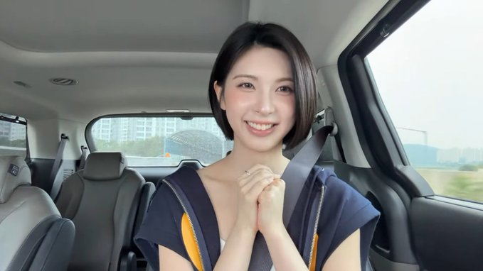 A smiling woman with short hair sitting in the back seat of a car, wearing a dark top and a seatbelt.