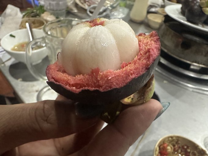 A hand holds a piece of exotic fruit showing white segmented flesh surrounded by a thick red rind, with a dark outer shell. A dining setting with various dishes is visible in the blurred background.