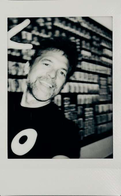A black and white polaroid-style photo of a smiling man making a peace sign in front of a bookshelf.