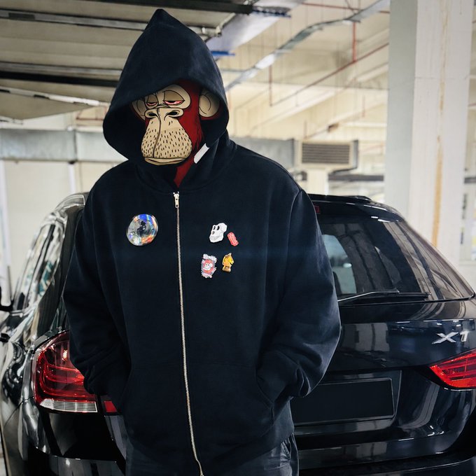 A person wearing a black hoodie with a monkey head mask and various pins/patches stands in a parking garage in front of a black BMW X1.
