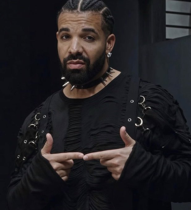 Portrait of the rapper Drake wearing an all-black outfit with a spiked choker and earrings, pointing his index fingers toward each other in a distinctive gesture against a dark background.