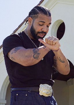 A man with braided hair and arm tattoos stands outdoors with his hands clasped together, wearing a black t-shirt and a large jeweled belt buckle. The image appears to depict Drake, who is projected to be the top Spotify US artist of 2026 according to Polymarket traders, reflecting his continued dominance in the music streaming industry.