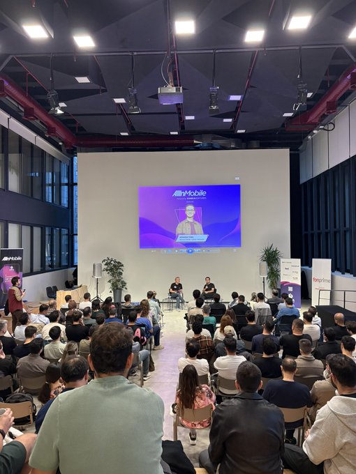 An audience sits in a large, industrial-style room watching two speakers on stage beneath a large screen displaying the 'Altr Mobile' logo and a virtual presenter.