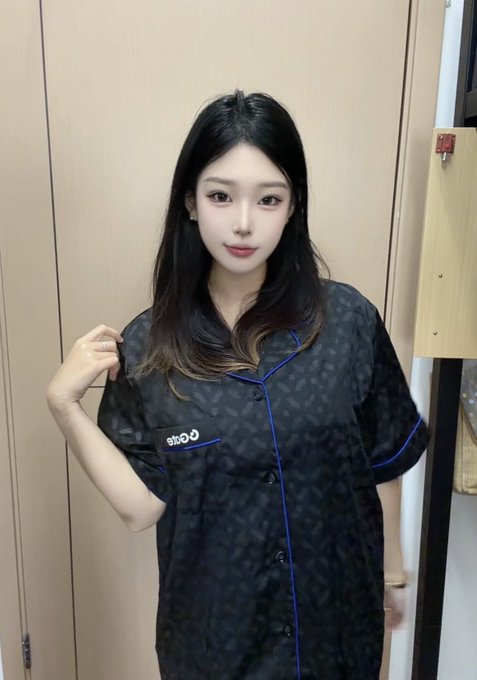 A young East Asian woman with long dark hair wearing a dark patterned salon smock with 'stbo-o' embroidered on it, standing in what appears to be a salon.
