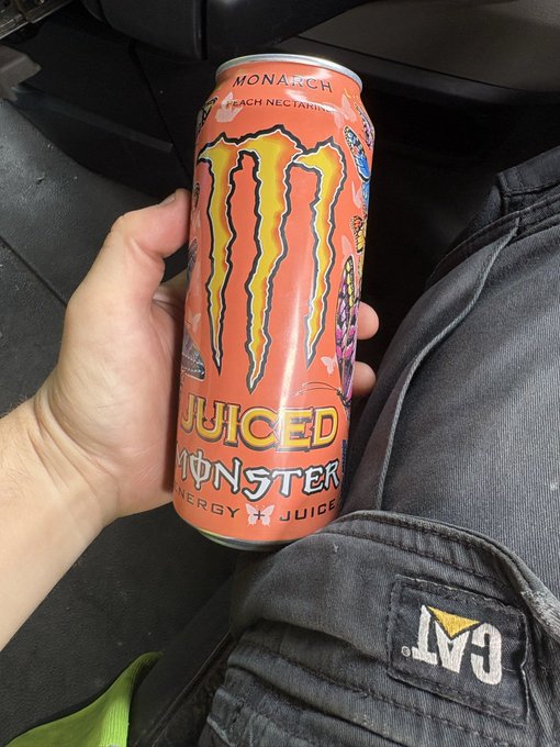 A hand holds an orange can of 'Juiced Monster Monarch Peach Nectarine' energy juice inside a vehicle, with a CAT-branded bag visible nearby.