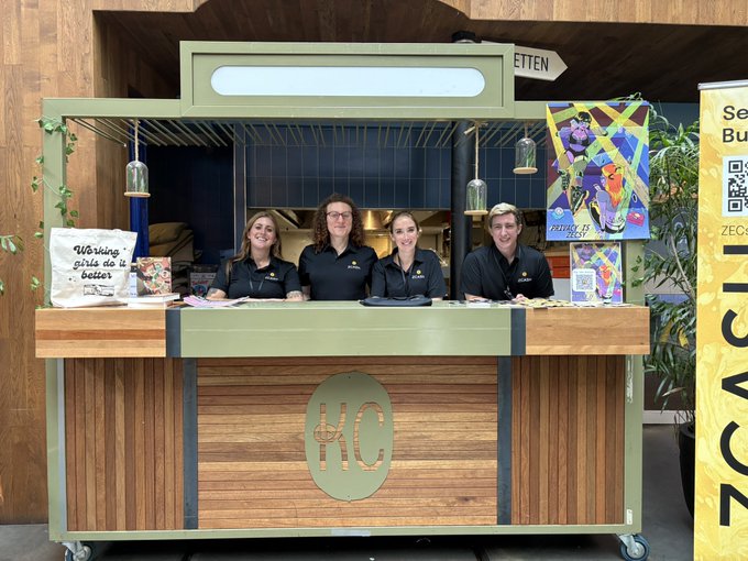 Four staff members stand behind a green and wood-paneled kiosk branded with "HC", displaying a sign that reads "Warung girls do it better".