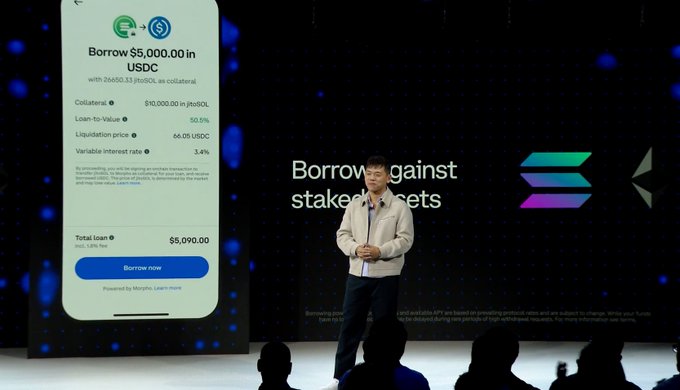 The image shows a man standing at a podium, giving a speech or presentation. Behind him, there's a large screen displaying information about a cryptocurrency called Sol token. The text on the screen reads "Borrow against your Staked SOL using $JitoSOL in Coinbase". This suggests that the speaker is discussing how to borrow against Sol token and use it as collateral for a cryptocurrency exchange.