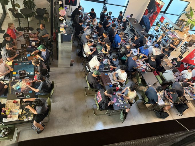 An overhead view of a crowded indoor event, possibly a gaming or trading card tournament, with many people seated at numbered tables engaged in gameplay or activities.