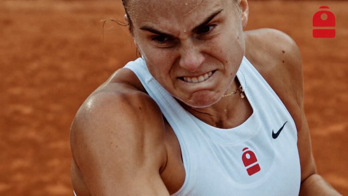 A close-up action shot of female tennis player Aryna Sabalenka on a clay court, looking intensely focused with a visible bite mark on her tongue, featuring a red logo in the upper right corner.