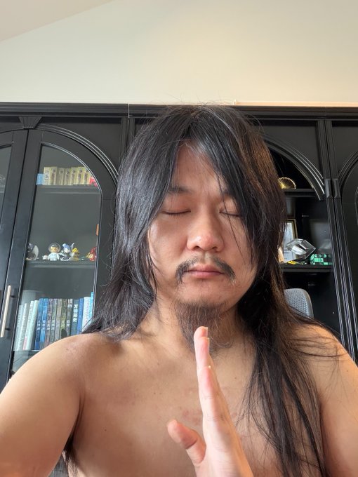 A shirtless man with long black hair and a beard poses with eyes closed and hands pressed together in a prayer gesture, standing in front of a dark bookshelf displaying figurines and books.