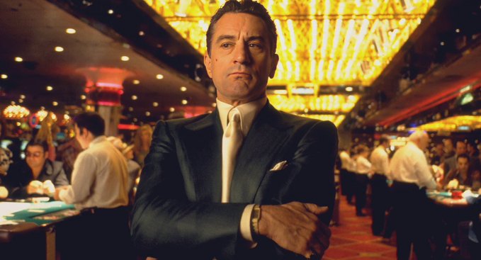 The image shows a sharply dressed man in a dark suit with a white tie, standing with crossed arms in an ornate, chandelier-lit casino—evoking the iconic casino boss aesthetic. The setting visually reinforces the post's gambling metaphor, suggesting strategic portfolio weighting in $TAO subnets where disciplined allocation (50% solid, 30% promising, 20% speculative) is the true edge, just as calculated bets separate a casino kingpin from a reckless gambler.