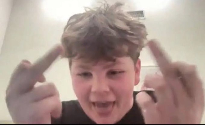 A young boy with messy brown hair makes peace signs with both fingers pointing toward his head while grinning excitedly at the camera.