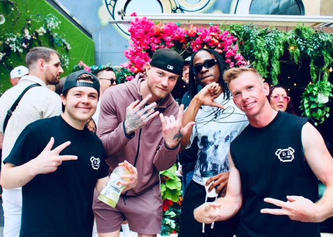 Photo of five young men posing together at what appears to be a social event, with flowers and greenery in the background; some are making hand gestures.