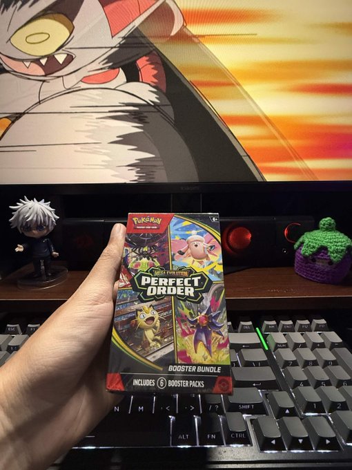 A hand holds a Pokémon TCG "Scarlet & Violet—Perfect Order" Booster Bundle (6 booster packs) featuring Raichu, Chansey, and Iron Crown, displayed in front of a keyboard and monitor showing a Meowscarada wallpaper, with anime figures visible on the desk—perfectly matching the post's excitement about picking up a booster bundle to open.