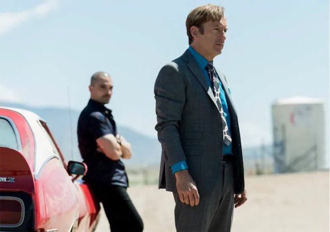 A man in a gray suit and bright blue shirt stands beside a red sports car in a sunlit outdoor scene, while another man is blurred in the background.