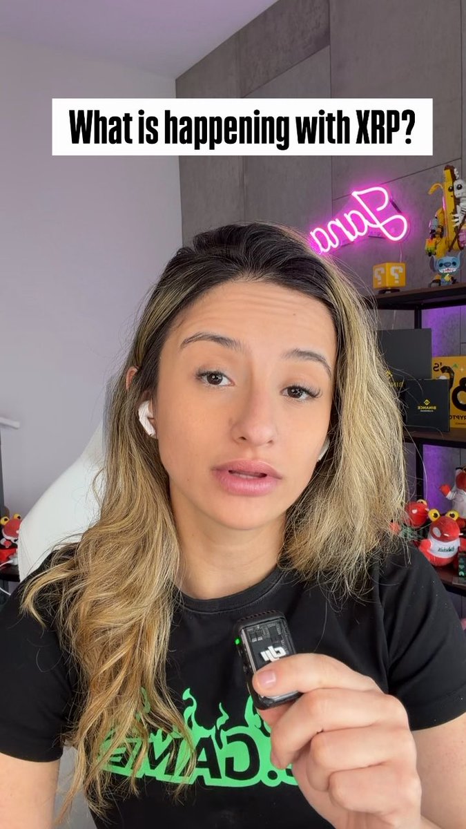 A woman wearing a black t-shirt holds a microphone and speaks directly to the camera, with on-screen text reading "What is happening with XRP?" above her in a room featuring a pink neon sign and various figurines.