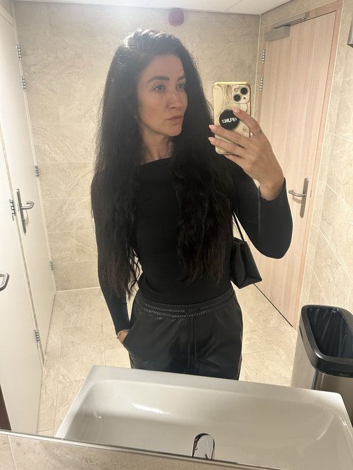 A woman with long dark hair poses for a mirror selfie in a bathroom wearing a black long-sleeve top and gray sweatpants, holding a phone with a PopSocket. The "GM" (Good Morning) post connects to her morning routine aesthetic while highlighting her optimism as an XRP holder, tying the casual self-portrait to crypto market resilience.
