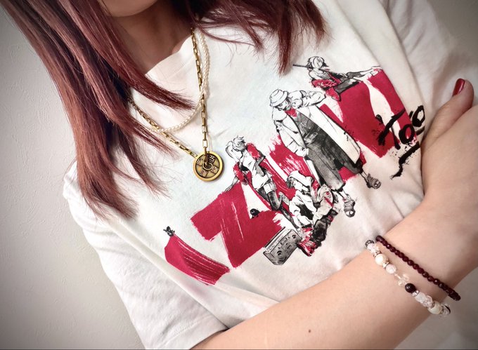The image features a woman wearing a white t-shirt with black text and a gold necklace featuring an Azuki logo, as well as two bracelets - one silver and one red - around her wrist. She has long brown hair tied up in a bun. The caption from the accompanying post reads: "Ohayo T-shirt season is here! An @Azuki T-shirt and an Azuki coin on a gold chain Favorites always make the day brighter Hope you have a wonderful day". The woman's outfit, accessories, and the presence of these elements in the image reflect a stylish and fashionable appearance.