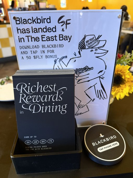 A promotional table display announces "Blackbird has landed in The East Bay" at Daytrip Counter, featuring a flyer inviting customers to download Blackbird for a "50 $FLY bonus," a "Richest Rewards in Dining" card highlighting up to 7X dining points when paying with Blackbird, and a circular "Tap Phone Here" NFC reader—showcasing the onchain payment integration connecting restaurants like the fried chicken spot to the Blackbird rewards ecosystem.