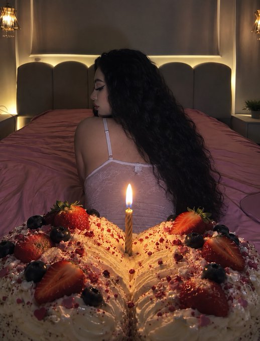 A woman with long dark hair sits on a bed behind an elaborate heart-shaped cake decorated with strawberries, blueberries, and sprinkles, topped with a single lit candle. The caption connects to her post about sponsoring cake at Token2049, with the lavish romantic presentation reflecting the "totally worth it" sentiment of her event sponsorship.