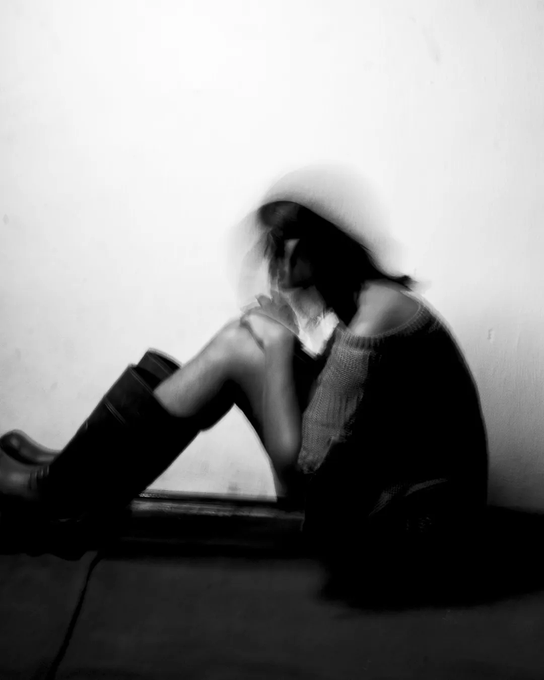 A black-and-white, motion-blurred photograph shows a person with long hair sitting on the floor with their knees pulled up, face obscured by their hair.