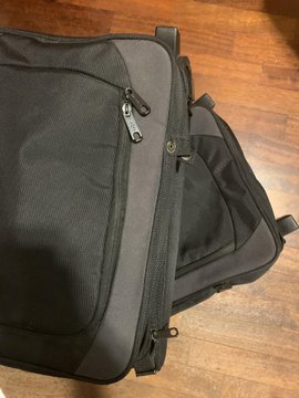 A close-up photograph of a black and gray fabric backpack or bag with multiple zippers, resting on a wooden floor.