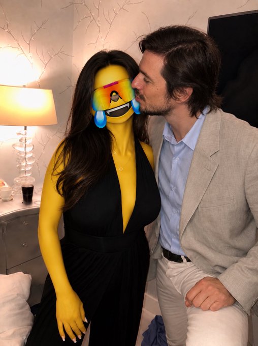 A photo of a man in a tan blazer kissing a woman in a black dress whose face is obscured by a laughing-crying emoji.