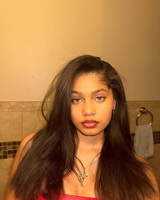 A young woman with long dark hair takes a bathroom selfie, wearing a red top and a cross necklace, with a towel ring visible behind her.