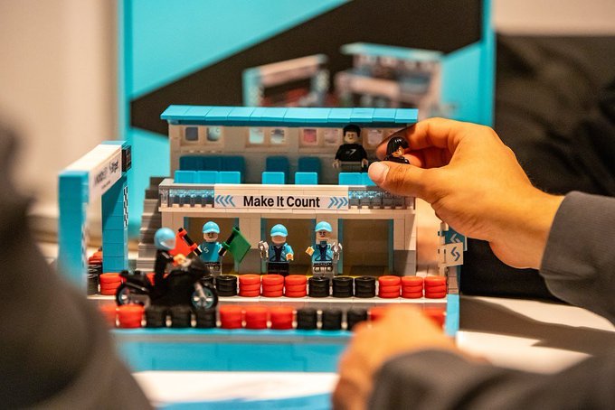 A close-up of a person's hand placing a Lego minifigure onto a detailed Lego stadium build labeled 'Make It Count', featuring small Lego police officers and red and black seats.