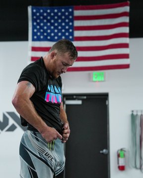 The image features a man wearing shorts with a graphic design, standing in front of an American flag. He appears to be preparing for a fight or practicing his moves, as he has his pants open and is holding a sports drink. The caption suggests that this could be related to a UFC event, possibly the one being presented by @Cryptocom.