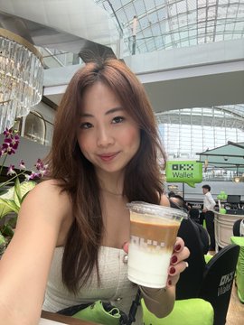 A selfie of a smiling young woman holding an iced latte inside what appears to be an airport or mall with DHL signage visible.