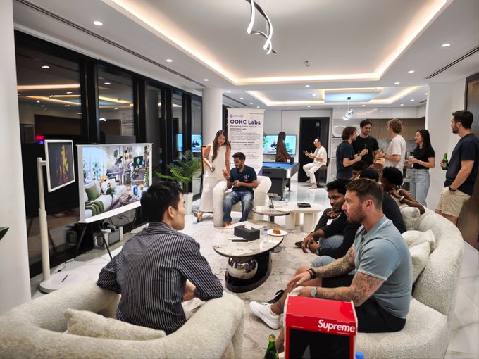A group of people gather in a modern living room with a large screen displaying a scene. A red shopping bag with 'Supreme' written on it sits in the foreground.