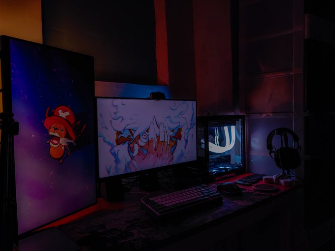 A dimly lit gaming setup featuring a monitor displaying an anime graphic of a hand making a gesture, alongside a vertical monitor showing a pirate-themed wallpaper, with a keyboard and headset on the desk.