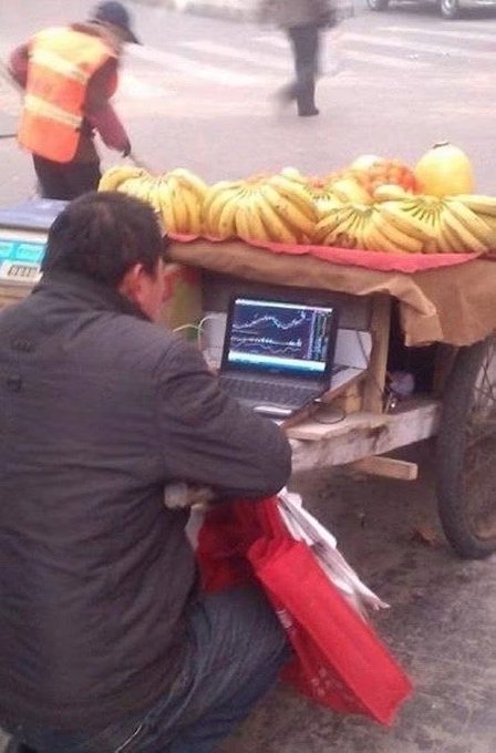A fruit vendor sits intently focused on his laptop displaying financial charts and graphs, while his cart of bananas and apples waits to be sold—a humorous commentary on misplaced priorities as a street sweeper works in the background.